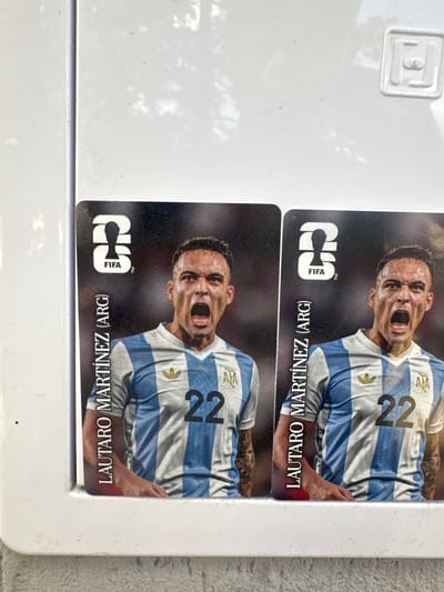Two collectible football cards featuring Lautaro Martínez of Argentina are displayed side-by-side. Each card shows a portrait of Martínez wearing the Argentine national team jersey, with his mouth open in a shout. The cards are placed on a white surface that appears to be a part of a white plastic cabinet or frame. In the background, a subtle embossed logo resembling a square with rounded corners and a horizontal line inside is visible on the white surface. The cards are positioned vertically, with text "LAUTARO MARTÍNEZ (ARG)" written vertically along the left edge of each card. The FIFA logo is also present on the top left of each card. The scene is brightly lit, suggesting an outdoor or well-lit indoor environment. The image does not contain any information about the location in Ovoshtnik, Bulgaria.
