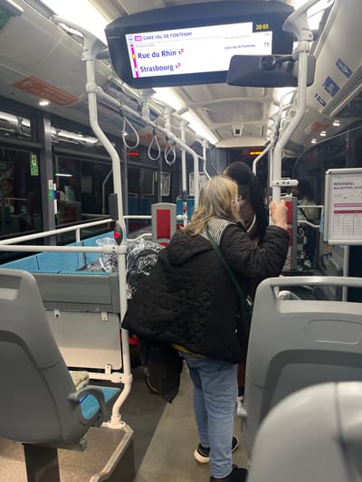 Inside a modern bus, a woman in a quilted jacket and blue jeans stands near a grey seat. An overhead display shows route information: line 86 with upcoming stops at "Rue du Rhin" and "Strasbourg," and the final destination "Gare Val de Fontenay." The current time is 20:03, with an estimated 15-minute arrival time at the destination. A sign about "Nouveau Navettes Sco" is visible above the woman. The bus interior features white handrails and red stop buttons.
