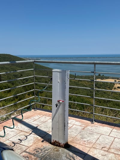 The image shows a partially weathered white water spigot with a red handle, situated on a tiled patio with a view of a body of water and lush green hills. A green coiled hose is attached to the spigot, extending to the left. A metal railing with horizontal bars runs along the edge of the patio, overlooking the landscape. The sky is clear and blue. The scene appears to be outdoors, possibly in a recreational area or a vantage point overlooking the Gargano National Park in Cagnano Varano, Italy. The sun casts shadows on the tiled ground.