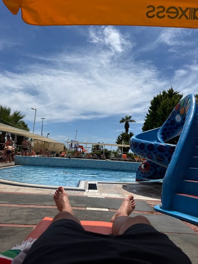 The image depicts a first-person perspective of a person relaxing on a lounge chair by a swimming pool. In the foreground, the person's legs and feet are visible, wearing black shorts. A striped towel is draped over the side of the lounge chair. The swimming pool is in the midground, with clear blue water and tiled edges. Several people are visible around the pool area, some sitting on lounge chairs and others standing. To the right of the pool, a bright blue water slide, designed to resemble a sea creature with eyes and patterns, is prominently featured. The background shows trees, including a palm tree, and a playground structure. The sky is blue with scattered white clouds. The overall scene suggests a resort or water park environment. The text "229Xi" is visible on the orange awning at the top of the image, which could be a logo or part of the establishment's branding. The location is identified as Ayvalık, Turkey.