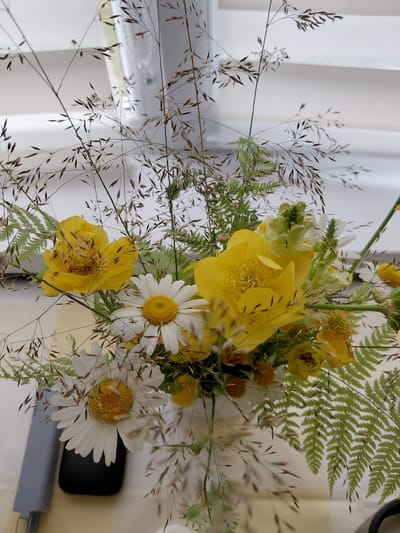 This is a close-up, vertical, eye-level shot of a floral arrangement. The arrangement is in a white vase, which is partially visible at the bottom center. The dominant flowers are bright yellow, with a ruffled texture, and white daisies with yellow centers. There are also some delicate green fern fronds and wispy, dried grass stalks interspersed throughout the bouquet. The background is blurred and appears to be an indoor setting, possibly near a window with white blinds. The overall impression is of a natural, rustic, and somewhat wild bouquet. There is no indication of the city of Chernyk, Ukraine, within the image itself.