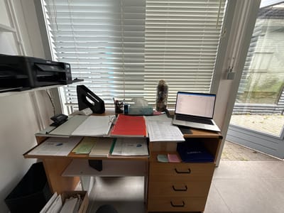 This image depicts a cluttered home office desk setup in Maldegem, Belgium, near a doorway leading outdoors. A black Epson printer is mounted on a shelf above the desk. The desk itself is made of light-colored wood and features a keyboard tray, a laptop, and various stacks of papers, folders, and office supplies. Notable items include a red binder, a tissue box, a decorative glass jar filled with dried materials, and a blue folder labeled "ALEKSIEV R.". A set of three wooden drawers is visible on the right side of the desk. A black trash bin is situated to the left of the desk, and a cardboard box filled with papers is on the floor below. The background shows a window with blinds and a partially open glass door revealing an outdoor area with paving stones and greenery. The overall impression is one of a functional, albeit disorganized, workspace.