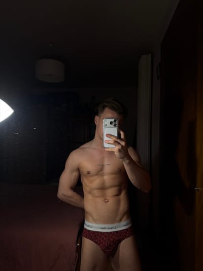 A shirtless man in red and black Calvin Klein underwear takes a mirror selfie, obscuring his face with his phone. He's slender and muscular, and there's a tattoo on his upper chest. The background is dimly lit, with a light fixture visible on the wall and a bed beneath the man. The mirror appears to be a bathroom mirror, capturing a portion of the man and reflecting the objects on the wall.