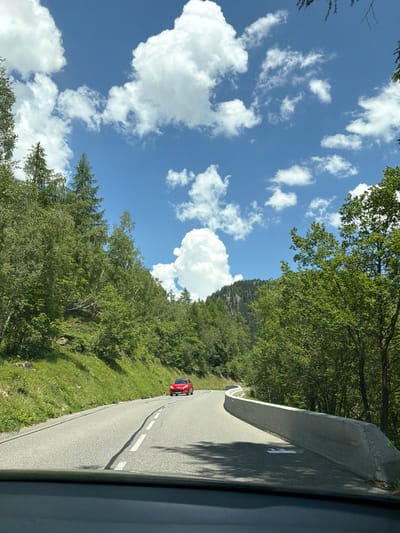 The image captures a view from a vehicle on a winding mountain road in Vallorcine, France. The road is paved and marked with white lines. A red car is driving ahead on the road. The road is bordered by a concrete barrier on the right and a grassy slope with trees on the left. The surrounding environment is lush with green trees and vegetation, suggesting a natural, mountainous area. The sky is blue with scattered white clouds. The scene is likely a scenic route within the French Alps.