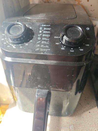 The image shows the top and front of a black DEXP air fryer, heavily coated in dust. The air fryer has two control knobs: one on the left with markings from 5 to 60, and another on the right with temperature markings from 80 to 200 degrees Celsius. A printed guide with icons and text in Russian is visible on the right side of the control panel, detailing cooking times and temperatures for various food items, such as pasta, french fries, chicken wings, and fish. The air fryer is placed on a countertop. The background is indistinct but appears to be an indoor environment. There is no visible indication of the city of Berdsk municipality, Russia, in the image itself.