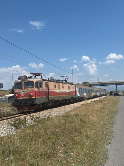 The image shows a train traveling on tracks under a bright blue sky with scattered white clouds. The train is red and beige with some graffiti on the side. In the background, there is a bridge or overpass and some vegetation. The location is identified as Mojanovići, Montenegro. There are no people clearly visible in the image. The train appears to be stationary or moving slowly. The environment is dry and sunny, suggesting a warm climate. The train tracks are surrounded by gravel and dry grass. A paved road runs parallel to the train tracks on the right side of the image.
