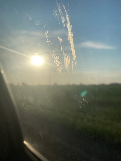The image appears to be taken from inside a vehicle looking out a smudged window. The sun is low in the sky, creating a bright glare and a hazy atmosphere, possibly indicating sunrise or sunset. The background shows a blurred landscape of green fields and a blue sky with some wispy clouds. There are prominent streaks and smudges on the window in the foreground, one of which appears to be a scratch. The left side of the image is dominated by the dark interior of the vehicle, with a bright reflection visible on what might be a dashboard or door panel.
Based on the provided information, the image was taken in the city of Rubtsovsk, Russia. The scene depicts a view from a vehicle in a rural or semi-rural setting, with the sun low in the sky. There are no people or specific objects clearly identifiable in the background, only the general environment. The smudges and scratches on the window are a key feature of the foreground.