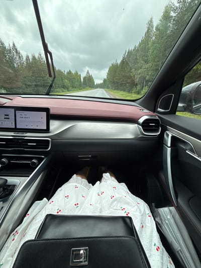 The image shows the interior of a car, looking out through the windshield at a road and a forest. The road appears wet, suggesting recent rain. The sky is overcast and cloudy. The dashboard of the car is visible, with a touchscreen infotainment system displaying radio information. A person's legs, wearing a white dress with a cherry pattern and yellow shoes, are visible in the passenger seat. A black handbag rests on the person's lap. The car is driving on a paved road that curves to the right, flanked by a dense forest of coniferous trees. The specific location in Krasnoyarsk, Russia cannot be determined from the image alone.
