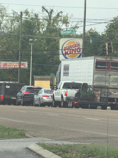 The image shows a street scene with several vehicles in traffic. In the background, there is a Burger King restaurant and a sign for "Riverport Rd 2900". There is also a sign for "Dickey's Pizzeria Italian Cuisine". In the foreground, there are several cars and trucks, including a white pickup truck pulling a trailer with a green piece of equipment on it. The sky is overcast. The scene appears to be in an urban or suburban area. The Burger King sign is a strong indicator that this is likely in a commercial district. The presence of traffic lights and multiple lanes of traffic suggest a busy intersection. The overall impression is a snapshot of everyday life on a road in a city.