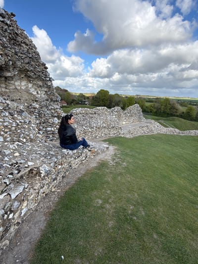 The image, when correctly oriented, depicts a serene outdoor scene featuring a lone person relaxing on a stone wall under a bright sky.
**Subjects:** The primary subject is a woman with long, dark hair, wearing a dark jacket and blue jeans. She is lying on her back, with her head resting on her hands, looking upwards.
**Objects:** The foreground is dominated by a robust, weathered stone wall, likely part of an ancient structure or ruins, constructed from numerous small, irregular stones, possibly flint, held together by mortar. Beyond the wall, there are expansive green grassy areas, possibly a field or hill. In the distance, a line of trees marks the horizon, and faint glimpses of what appear to be rural buildings or a small settlement can be seen. The sky is filled with large, white, fluffy cumulus clouds interspersed with patches of clear blue sky.
**Scene/Setting:** The setting is an outdoor historical or natural site, characterized by ancient stonework and open green landscapes. Given the location context of King's Lynn and West Norfolk in the United Kingdom, this could be an area with historical fortifications, castle ruins, or an ancient earthwork like a hillfort.
**Activity or event taking place:** The woman is engaged in a moment of quiet relaxation or contemplation, lying down and gazing at the sky.
**Notable details:**
* **Time of day:** It is daytime, indicated by the bright lighting and clear visibility.
* **Weather:** The weather appears fair, with a mix of sunshine and significant cloud cover.
* **Location cues:** The ancient-looking stone wall and rolling green terrain suggest a site of historical or archaeological interest.
* **Emotions:** The woman's relaxed posture and upward gaze convey a sense of calm and peaceful observation.
* **Visible text:** There is no visible text in the image.