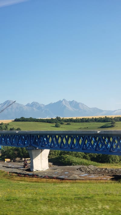 The image captures a scenic view of the High Tatras mountains in the background, with a bright blue sky above. In the foreground, a large blue truss bridge under construction is visible, supported by a concrete pillar. The bridge spans across a landscape that includes green fields, trees, and a body of water. The area around the bridge appears to be a construction site, with piles of gravel, construction vehicles, and what looks like red piping on the ground. In the mid-ground, there are more fields and trees, with the majestic mountain range forming a dramatic backdrop. The overall impression is one of natural beauty juxtaposed with ongoing infrastructure development in the vicinity of Gánovce, Slovakia.