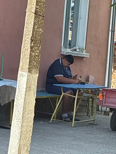 An adult male with dark hair, wearing a navy blue polo shirt with white and blue striped trim and navy blue shorts, is seated on a blue bench at a blue-topped table. He is leaning forward, focused on a smartphone held in both hands. White socks and dark sandals are visible on his feet. A small, pink object rests on the table to his right. The background features a building with an orange-brown stucco-like wall. A vertical window with a white frame is present in the wall, with several items on its sill, including a crumpled white object and a small green glass. To the left, a portion of another table covered with a white lace tablecloth and a green metal pole are visible. To the right, part of a red trailer with an orange circular reflector and a black tire is present. A prominent, rough-textured, light-colored concrete pillar is in the left foreground, partially obscuring the view, with a dark wire wrapped around it. The ground surface is light grey asphalt or concrete. The scene is located in Radievo, Bulgaria.