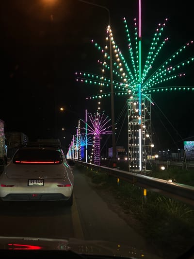The image shows a street scene at night, likely in Lang Suan, Thailand, adorned with colorful lights. The primary subjects are various light installations, shaped like exploding fireworks or starbursts, made of numerous small, multicolored LED bulbs. These lights, predominantly green, pink, and purple, are strung along poles and scaffolding, creating a festive and vibrant atmosphere.

In the foreground, the rear of a white car is visible, facing away from the viewer. The car has its taillights illuminated, showing a red glow. A license plate with Thai characters and the number "ขง 8225" is clearly visible. Beyond the car, the road continues, bordered by a grassy embankment and a metal railing.

The scene appears to be part of a nighttime event or festival, possibly a celebration or a roadside display. The darkness of the night sky contrasts sharply with the bright, artificial illumination of the lights, which dominate the visual space. There are also some larger, more industrial-looking structures with more lights, suggesting a larger display or fairground. The overall mood is festive and visually striking due to the abundance of colorful lights against the dark backdrop. No people are clearly visible in the image. The weather appears to be clear, allowing for the bright display of lights.