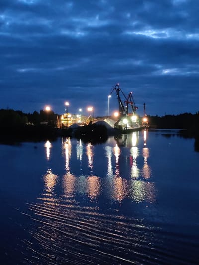The image depicts a body of water at dusk or night, reflecting lights from a construction or industrial site on its far bank. The site features crane-like structures and brightly lit buildings, possibly a quarry or factory. The water surface exhibits ripples, indicating movement. On either side of the central illuminated area, darker silhouettes of trees suggest a wooded shoreline. This scene is located in Александровское, Russia.