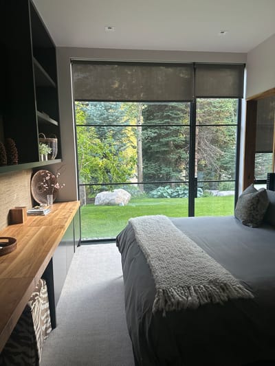 The image depicts an interior space, likely a bedroom, with a view of an outdoor landscape, located in Aspen, United States. The room features a large, floor-to-ceiling window with black frames that divide the glass into multiple panes. Dark grey roller blinds are partially lowered over the top sections of the window.
To the left, a dark-colored built-in shelving unit contains decorative items, including two pinecones, a small basket with a white container and green stems, and another small woven basket. Below the shelving, a light brown wooden countertop/desk area rests on dark grey cabinetry. A textured light brown wall covering serves as a backsplash. On the countertop, a dark woven dish leans against the wall, alongside a clear glass vase with pink floral stems, a small wooden box, and a stack of flat objects. Beneath the countertop, a basket or bag with a white and brown animal print is visible. The floor is covered in a light grey patterned carpet.
In the foreground, a bed with dark grey bedding is positioned parallel to the window. A light grey, chunky knit throw blanket with fringe is laid across the foot of the bed. Two pillows, one with a subtle patterned design, are visible at the head of the bed. The ceiling is white and includes recessed lighting fixtures.
The outdoor environment visible through the window consists of a well-maintained green lawn. A large, light-colored boulder is prominently situated on the lawn. Beyond the lawn, a dense array of mature trees, including both conifers and deciduous trees, forms a natural backdrop. No individuals are present in the scene.