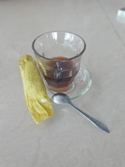 This image captures a close-up view of a glass of dark liquid, likely coffee, placed on a small glass saucer. Beside the glass, a crumpled piece of bright yellow material, possibly wrapper or a small snack, rests on a light-colored, speckled surface, resembling a tiled countertop. A silver spoon lies diagonally across the scene, its tip pointing towards the bottom right corner. The lighting suggests it is daytime, with reflections visible on the glass surfaces. The scene appears to be set indoors, likely in a casual environment such as a kitchen or a cafe in Keudeteupinpunti, Indonesia. There is no visible text in the image.