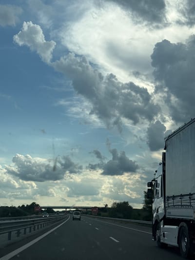 The image depicts a view from a vehicle traveling on a highway. The sky is filled with a dramatic mix of dark, heavy clouds and patches of bright blue sky. In the foreground, a white truck is partially visible on the right side of the frame, traveling in the same direction. The highway stretches ahead, with other vehicles, including a white van and a car, visible in the distance. The road is bordered by safety barriers and greenery on the left. An overpass is visible in the far distance. The overall impression is one of a journey under a dynamic and changing sky. The location is specified as the city of Yazdach, Bulgaria.