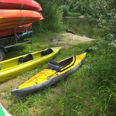 The image depicts a riverside scene in Хотылёво, Russia. In the foreground, two kayaks are resting on grassy ground. One is a bright yellow kayak with a black seat and paddle. Next to it is a lime green kayak, also with a black seat. Behind these, a trailer is visible, loaded with several red and orange kayaks stacked on top of each other. A dark green kayak is also on the trailer. The river, with calm, green water, is visible in the background, bordered by lush green trees and foliage. A sandy bank with a fallen log is present near the water's edge. The overall setting suggests an outdoor recreational area by a river.