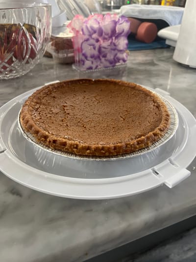 A round, golden-brown pie rests in an aluminum pan, topped with a crimped crust. The pie is centered on a white plastic serving dish with a clear plastic cover. To the left are a bouquet of purple and white flowers in a plastic container and a cut-glass bowl filled with fruit. The scene is set on a gray marble countertop.