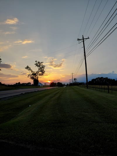 A rural road is seen at sunset in Lancaster, United States of America. The sun is setting behind a lone tree and a white house, casting a warm glow across the sky and illuminating the clouds with shades of orange and yellow. The road is paved and has a white line marking the edge of the lane. On the right side of the road, a grassy embankment slopes upwards towards a fence and a line of trees. A utility pole with power lines stretches across the sky, a common sight in rural areas. The grass is green and well-maintained, suggesting it's likely a well-trafficked area, perhaps near a farm or residential property. The overall scene is peaceful and serene, capturing the beauty of a rural sunset.