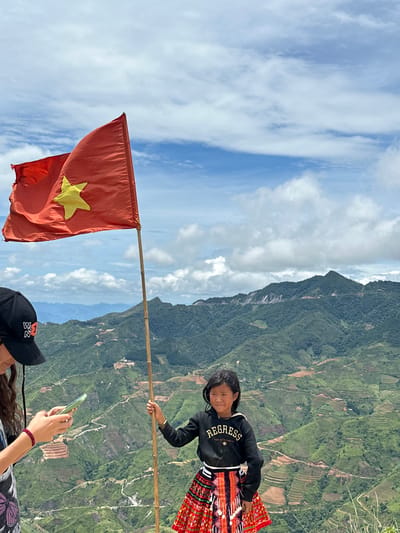Two individuals are positioned on a scenic overlook, with a Vietnamese flag flying prominently. A younger girl, wearing a black hoodie and a vibrant red and black patterned skirt, stands with her left hand grasping a bamboo pole that supports the flag. To her left, an individual wearing a black baseball cap and a dark shirt is looking at a mobile phone held in their left hand. The background showcases lush, green mountains with terraced fields and winding roads, indicating a rural mountainous region, likely in the vicinity of Bắc Yên, Vietnam. The sky above is partly cloudy, with white clouds scattered across a blue expanse. The scene appears to be a moment captured during an outdoor excursion.