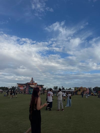 The image displays a large outdoor event situated on an expansive green grassy field under a partly cloudy sky, with a faint rainbow visible in the upper left quadrant. The location is an open area within the city of Martínez, Argentina. In the immediate foreground, a female with long, dark reddish-brown hair is visible from the back-left, wearing a dark top and a dark long skirt or dress, holding a white smartphone in her right hand. Numerous individuals are scattered across the midground and background, either standing or walking. Notable figures include a male in a white baseball jersey with the number "32" on its back, and another male in a light-colored striped shirt and light grey pants, both seen from behind in the midground.
In the background, a large stage structure features a prominent screen displaying text, partially obscured by a tall, multi-colored inflatable or sculptural installation resembling stacked shapes. Text on the screen appears to include "70,000 HECTAREAS AFECTADAS". To the right of the main stage, another significant, multi-colored inflatable structure is present. Tall metal towers, presumably for lighting or sound, are also visible in the distant background. The horizon line indicates a treed area behind the event structures.