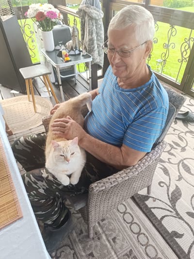 An elderly man with white hair and glasses sits in a wicker chair on a covered porch, holding a light-colored cat on his lap. He is wearing a blue and white striped t-shirt and camouflage pants. The cat is looking towards the viewer with its eyes partially closed. In the background, a table holds a vase of flowers, a thermos, and some food items. A camouflage jacket hangs on a railing behind the table. The porch has a wooden floor and is furnished with a rug. The scene appears to be in the village of Pliusnino, Russia, indicated by the presence of outdoor furniture and a natural, green outdoor environment visible through the porch railings.