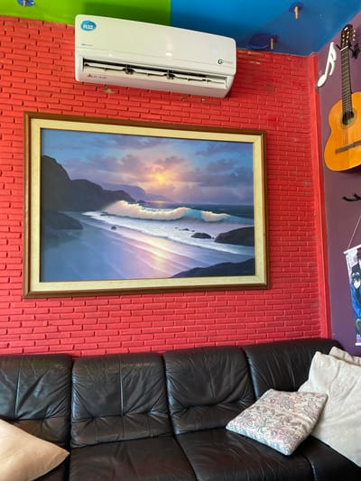 The image depicts an interior scene with a black leather sofa in the foreground, adorned with two pillows. Behind the sofa, a vibrant red brick wall serves as a backdrop. Mounted on this wall is a framed oil painting of a serene beach landscape with crashing waves and a sunset. Above the painting, a white air conditioning unit is installed. To the right of the air conditioner, an acoustic guitar is hung on the wall, along with a musical note-shaped decoration. Further to the right, a banner featuring anime characters is suspended. The ceiling is painted blue and has some exposed wiring and fixtures. The overall impression is that of a cozy, artistically decorated room, possibly a living room or lounge area. The location specified is Bang Sare, Thailand.