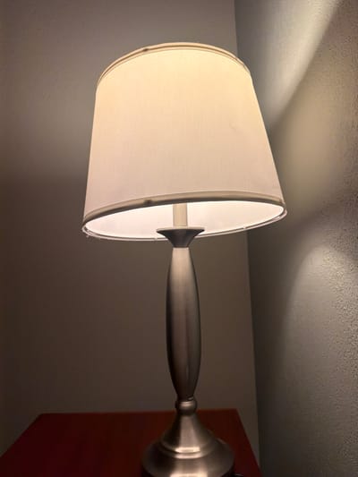 An indoor scene in Dover, United States of America, depicts a lit table lamp positioned on a dark reddish-brown wooden surface. The lamp consists of a brushed metal base and an elongated, tapering stem. A white fabric drum lampshade, internally illuminated, rests atop the stem. The lampshade's upper and lower rims are trimmed, exhibiting minor dark blemishes and slight fraying on a section of the lower rim. The lamp casts light onto a textured wall on the right and an adjoining smoother wall on the left. No individuals or specific actions are present within the frame.