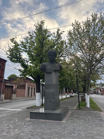 The image captures a view of a street scene in the city of Akhieli, Georgia. A stone bust of a man stands prominently on a plinth. The monument is situated along a sidewalk paved with interlocking stones. Behind the monument and sidewalk are trees, lining the pedestrian walkway, some of which are painted white at the base. A brick building is adjacent to the walkway. In the distance, other buildings can be seen along the road. The sky is visible above the buildings and trees.