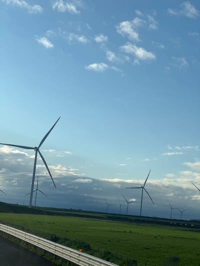 The image displays a rural landscape near Carapelle, Italy, under a blue sky with scattered white clouds. Multiple wind turbines are the primary subjects, distributed across the middle ground and background. One prominent, three-bladed wind turbine with red-tipped blades is positioned in the mid-left foreground. Additional three-bladed turbines are visible further back, becoming progressively smaller and less distinct towards the horizon, where a band of darker, extensive clouds partially obscures their bases. The foreground consists of vibrant green fields. In the immediate lower-left, a multi-segmented, reflective metal guardrail with orange markers runs diagonally, separating a dark surface, likely a road or shoulder, from the green field. No individuals, animals, or active movements are depicted.