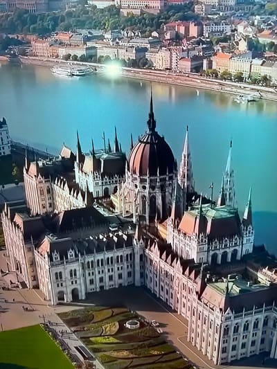 This media file is an aerial, wide-angle photograph of a city with a river flowing through it. The image is oriented vertically, giving it an unusual perspective. In the foreground, the prominent subject is a large, ornate building with a domed roof and multiple spires, suggesting a landmark or significant architectural structure. The building exhibits intricate detailing and a light-colored facade.

The river, a wide expanse of blue water, is visible to the right of the building. Along the riverbank on the left side of the image, there is a dense urban area with many smaller buildings, characterized by red-tiled roofs and a mix of light and dark facades. Trees are interspersed among the buildings, indicating a green urban landscape. The overall impression is of a historical European city, likely situated on a river.

The lighting in the image suggests it was taken during the daytime, with a bright reflection on the water indicating sunlight. The sky is a clear, light blue. There are no people or specific activities clearly depicted, focusing instead on the architectural and environmental elements of the scene. The image lacks any visible text. The provided location context, "Ləcət, Azerbaijan," does not appear to align with the visual cues of the architecture, which strongly resemble buildings found in cities like Budapest, Hungary, or other Central European capitals.