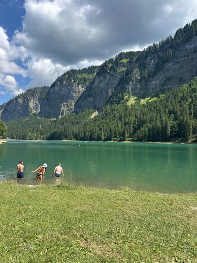 The image displays Lac de Montriond in Montriond, France, under a sky with mixed blue patches and substantial grey and white clouds. In the foreground, a green grassy bank slopes down to the water's edge. Three individuals are wading in the emerald-green lake near the shore, facing away from the viewer. From left to right, a male wearing dark swim shorts, a female in a dark bikini and white wide-brimmed hat, and another male in dark swim shorts are partially submerged, appearing to enter or stand in the water. The background is dominated by large, steep mountains densely covered with coniferous trees, featuring visible rock faces and natural slopes. The mountains reflect faintly on the calm lake surface. Distant yellow buoys and faint figures of other individuals or watercraft are visible further out on the lake.