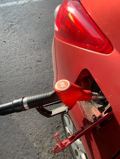 The image shows a close-up, angled view of a red car being refueled at a gas station. A bright orange fuel nozzle with "95" displayed on it is inserted into the car's fuel tank. The nozzle is connected to a black hose. The car's rear taillight is visible, as is a portion of a silver wheel. The ground appears to be asphalt. The scene is set outdoors, under bright sunlight. The location is stated to be Belogorsk, Russia.