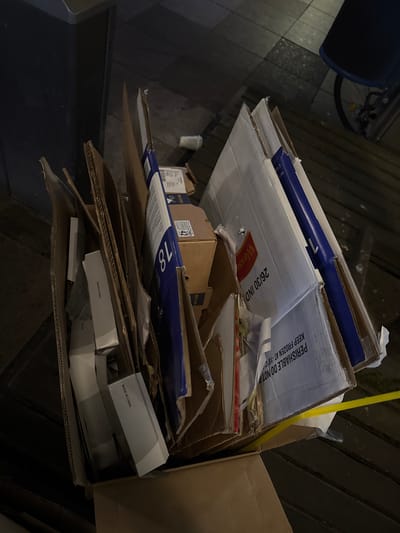 The image shows a pile of cardboard boxes and other packaging materials, suggesting a scene of refuse disposal or recycling. The boxes are of various sizes and designs, with some bearing labels and branding. One prominent box is a large, white one with blue trim, and it has text on it that includes "WESSO" and "26/30 IND." Another box has the number "18" in large blue digits. Within the pile, smaller white boxes are visible, some with the text "MADE IN CHINA" printed on them. A yellow strap is also visible, possibly used to secure the contents of the pile. The scene appears to be outdoors, possibly on a street or sidewalk, given the presence of paving stones in the background. The overall impression is one of discarded materials waiting to be collected. There are no people visible in the image, and the focus is entirely on the collection of waste. The location is identified as Duisburg, Germany.
