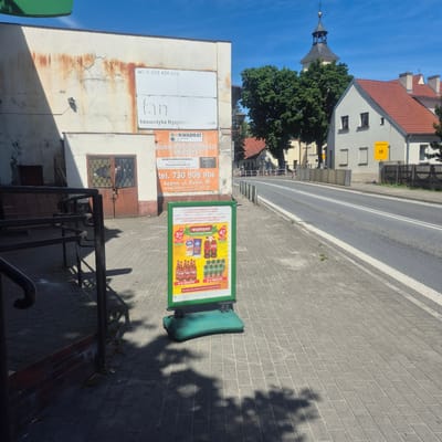 The image shows a street scene in Bralin, Poland, on a sunny day. In the foreground, a green A-frame sign is placed on a paved sidewalk. The sign advertises a "WEEKEND" promotion with discounts on beer and Coca-Cola. Various bottles of beer and cans of soda are depicted.

To the left of the sign, the exterior of a building is visible, showing a distressed facade with cracks and staining. A set of metal railings and a brick wall are also present. Further back, a building with a metal door and barred windows can be seen.

Across the street, residential buildings with red-tiled roofs are visible, along with a church tower in the background. A yellow sign with figures of children is mounted on a fence. The street is paved with asphalt and marked with white lines. The sky is clear and blue. The scene appears to be in a commercial or mixed-use area of the town.