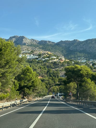 The image shows a road in Altea, Spain, on a sunny day. The road is paved and has white lines marking the lanes. A guardrail runs along the side of the road, separating it from a steep, tree-covered hillside. Several vehicles are visible on the road, including a truck and a car.
The hillside is dotted with buildings, suggesting a town or village is nestled into the landscape. The architecture appears modern, with white and light-colored structures, some of which are built into the hillside. Dense green trees cover much of the terrain, with a few taller trees closer to the road. In the distance, a large, rocky mountain looms over the scene. The sky is a clear blue with a few wispy clouds. The overall impression is a scenic drive through a Mediterranean landscape. The time of day is likely midday, given the bright sunlight and distinct shadows cast by the guardrail and trees.