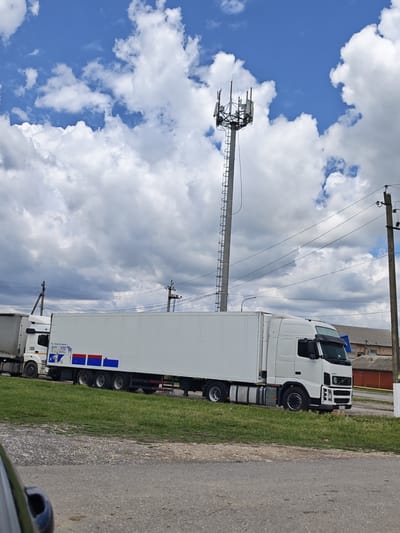 A white semi-trailer truck is parked on the side of a road with a cell tower in the background. The truck has a long white cargo trailer and a white cab with a Volvo logo. The truck is on a grassy area next to a paved road. The cell tower is a tall metal structure with antennas at the top, and it is silhouetted against a blue sky with scattered white clouds. There are power lines running across the image. The overall scene appears to be in an industrial or semi-rural area. The license plate on the truck is partially visible and appears to be in Cyrillic script.