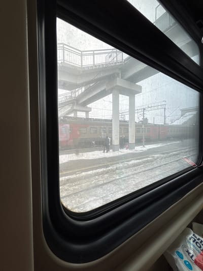 The image captures a detailed view from inside a train car looking out a window at a railway station in Barabinsk, Russia, under overcast winter conditions with light precipitation. The window frame, a dark rubber seal, and a light-colored interior wall are prominent in the immediate foreground. Outside, snow and slush cover the ground, tracks, and portions of the platform.

A stationary train, comprising multiple cars painted red and beige, occupies the tracks directly adjacent to the platform. On the snow-dusted platform, approximately five individuals are observed. To the left, two figures stand near a train car, one with their back to the viewer, facing the train. Further to the right, a small group of three people is gathered on the platform, with a pink bag visible near them, suggesting luggage.

In the background, a large concrete pedestrian overpass with metal railings spans the railway area, supported by thick concrete pillars. Overhead electrical lines and railway catenary systems are visible beneath and around the overpass. The sky appears bright but hazy, consistent with the observed weather. A white plastic bag with blue text, possibly containing disposable wipes, is visible inside the train car in the lower right corner.