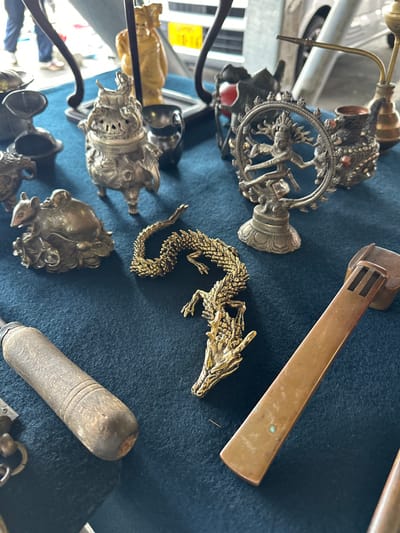 This image presents a close-up view of various antique or vintage objects displayed on a dark blue felt surface at an outdoor market in Shinagawa, Japan.

In the center foreground, a detailed, textured brass-colored coiled dragon statuette lies horizontally. To its immediate left, a bronze-toned statuette of a rat or mouse is depicted holding several round objects. Further left, extending from the lower corner, is the dark wooden handle of a tool, possibly a chisel, with a metal ferrule. Above the rat statuette, a silver-toned, ornate, three-legged incense burner with a lid stands prominently. A small, dark, plain bowl is positioned to the right of the incense burner.

In the midground, a silver-toned statuette of the Hindu deity Shiva Nataraja, dancing within a circular frame, is centrally placed to the right of the dragon. Adjacent to the Nataraja figure, a small, ornate metal pipe with copper-colored accents and minor red embellishments is visible. In the upper-midground, behind the incense burner, a light-colored, possibly wooden or ivory-toned statuette of a robed figure, resembling a laughing Buddha, stands on a small base.

The background is significantly blurred, indicating an outdoor setting. Visible elements include portions of a white vehicle, specifically a fender, and a yellow Japanese license plate displaying "し 12-42". The lower legs of a person wearing blue pants and light-colored shoes are also faintly discernible, confirming a public, open-air environment. No explicit actions or interactions are occurring; the scene is a static display of merchandise.