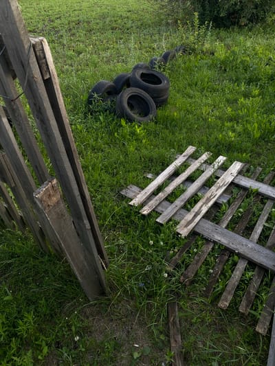 This is a high-angle, medium shot of an outdoor scene in Oktyabr'skii, Russia, with a focus on weathered wooden fencing and discarded tires amidst overgrown green grass and clover. The wooden fence is constructed from rough, aged planks, some of which are broken or detached, indicating disrepair. A section of the fence is laid horizontally on the ground, appearing to be part of a dismantled structure.
In the left portion of the image, a stack of five black tires is partially submerged in the tall grass. The tires are worn, suggesting they have been used and discarded. The surrounding area is densely covered with green grass, interspersed with small white flowers, likely clover. Towards the top left, dense foliage and greenery are visible, hinting at the presence of trees or bushes.
The scene appears to be set during the daytime, with natural light illuminating the area. There are no people or animals visible. The overall impression is one of neglect and abandonment, with natural elements reclaiming man-made objects. The wooden materials suggest a rural or semi-rural setting, possibly a yard or an abandoned lot. No text is visible in the image.
