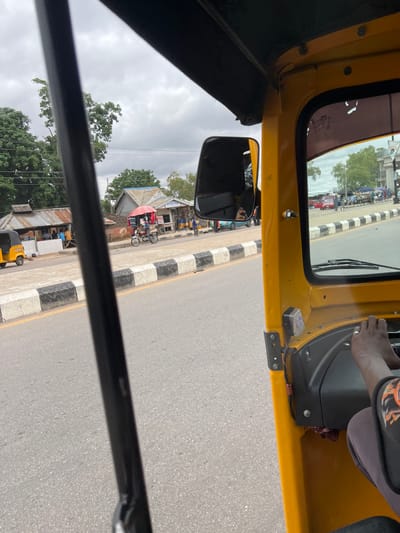 An exterior, medium shot captures a view from inside a yellow three-wheeled auto-rickshaw. The driver's hands are visible on the steering wheel, and their torso is partially visible. The vehicle is on a paved road, bordered by a sidewalk with black and white striped curbs.

To the left of the road, a small marketplace is set up with several stalls. One stall features a corrugated metal roof and is supported by wooden posts, with a few people standing and sitting outside. Another stall has a red awning and a motorcycle parked in front of it. Further down the road, more stalls and a few cars are visible.

The environment appears to be a populated area in Kporva, Nigeria, with a mixture of buildings, vehicles, and pedestrian activity. The sky is overcast, suggesting a cloudy day. The overall scene depicts a typical street view within a bustling Nigerian town.