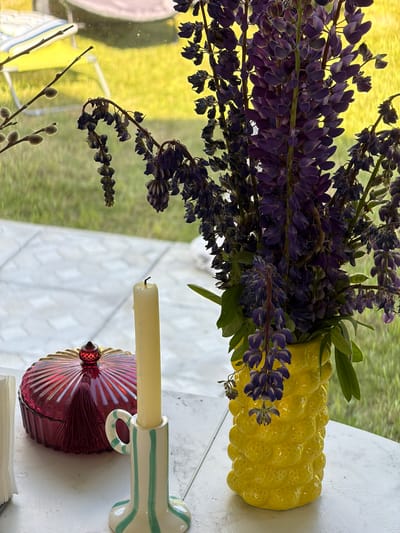 The image depicts a still life arrangement on a white table with a tiled outdoor background. In the foreground, a tall, unlit cream-colored candle stands in a small, striped ceramic candle holder with a light green and white stripe pattern and a loop handle. To the left of the candle is a decorative, deep red glass dish with a fluted lid and a small, faceted red finial on top. A dark red tassel hangs from the lid.
Behind these objects, a bouquet of dark purple lupine flowers is arranged in a bright yellow, textured ceramic vase that resembles a pineapple. The flowers are mostly in bloom, with some withered blossoms visible. Green leaves are interspersed with the flowers.
Through a window or glass partition in the background, a grassy area with blurred green and yellow tones is visible. A yellow chair with a striped cushion is also discernibly out of focus. The tiled ground in the background suggests an outdoor patio or walkway. There are no people or animals present in the image. The setting appears to be a domestic outdoor space, potentially a patio or balcony, in or near Алешкино Дп, Russia.
