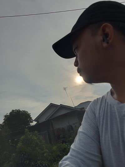 The image, taken in Teluk Kenidai, Indonesia, shows a close-up of a person wearing a black baseball cap and a light-colored t-shirt, with a dark earbud in their right ear. The person is looking to the left, away from the camera. The background features a hazy sky with the sun visible and appearing somewhat diffused. Below the sky, there are residential buildings with visible roofs and a tree. The scene is characterized by natural light, possibly from a late afternoon or early morning setting. The perspective suggests a selfie or a photo taken from a slightly elevated position.