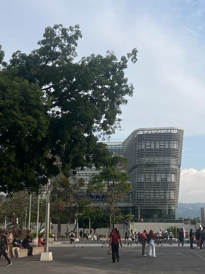 The image captures a scene in San Salvador, El Salvador, with a modern building featuring a curved facade with horizontal lines, likely an office or commercial building. The building is surrounded by trees and greenery. In the foreground, a plaza is visible, populated by people walking and sitting, with pigeons present on the ground. The sky is overcast.