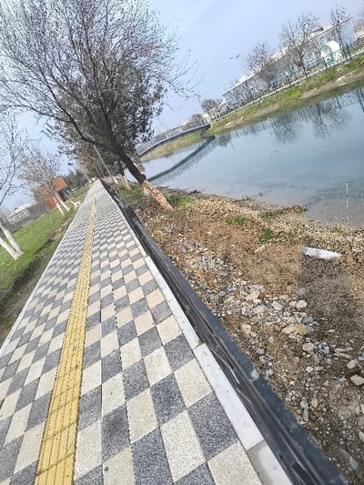 The image depicts an outdoor urban scene in Pakhtakor, Uzbekistan, under an overcast sky. A newly paved pedestrian walkway extends from the foreground into the mid-distance on the left, featuring a checkerboard pattern of light grey and dark grey rhombus-shaped tiles. A yellow tactile paving strip runs longitudinally down the center of this walkway. Adjacent to the walkway on its right, a dark, low barrier separates it from a sloping dirt and gravel embankment that descends towards a calm body of water, likely a canal or river. The embankment contains scattered rocks, bare earth, and sparse patches of green vegetation.
In the mid-ground, a light-colored arch pedestrian bridge spans the water, with its reflection visible on the water's surface. On the far bank, beyond the water and bridge, several light-colored buildings with dark roofs and multiple leafless trees are present. A small, light green structure is also visible near the buildings. A row of bare deciduous trees with some white-painted bases lines the left side of the walkway. No individuals or specific actions are discernible within the frame.