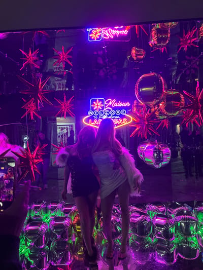 The image shows two women posing in a mirrored room decorated with balloons and neon signs. One neon sign reads "Maison Lacome" and another reads "Fabulous Las Vegas." The balloons are arranged in clusters near the top of the room and near the right side. A hand holding a smartphone is visible in the bottom right corner, suggesting someone is taking a photo. The environment is bathed in purple and green lighting. It is not possible to determine the location in Ezeiza, Argentina from this image.