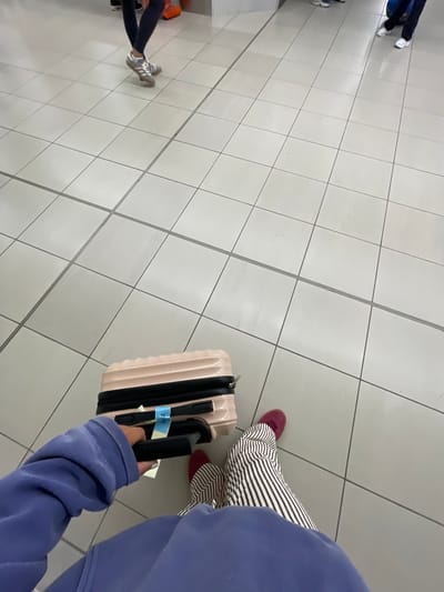 The image, captured from a first-person, downward-looking perspective, depicts a scene likely within Gatwick Airport (LGW) in Crawley, United Kingdom. In the foreground, the individual's right arm, clad in a blue long-sleeved garment (possibly a hoodie), extends to grip the black, extended handle of a light beige or pink hardshell rolling suitcase. A light blue luggage tag, possibly bearing the inscription "AGE," is attached to the handle. Below, the individual's lower legs are visible, wearing white and dark striped full-length pants, and burgundy-colored flat shoes.
The floor is covered with large, light-colored square tiles with dark grey grout lines, indicating an indoor public space. In the midground and background, several other individuals are partially visible, identified by their legs and feet. One person wears dark leggings and grey and white athletic shoes. Another person wears dark trousers and white athletic shoes. An additional pair of feet is visible further back, near an orange object on the floor. All visible individuals appear to be standing or moving slowly, consistent with a queue or busy thoroughfare within a travel terminal. The overall scene conveys a sense of waiting or transit.