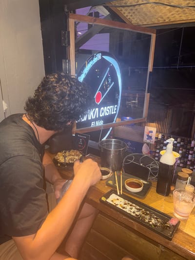 A medium shot captures a seated individual with curly brown hair, wearing a black t-shirt, facing a television displaying the logo "RIKA CASTLE El Nido." The person appears to be dining, with a partially consumed dish in front of them and a utensil in their hand. On the right side of the image, a wooden table holds various items, including a small bowl with a dark liquid, an empty sushi tray, a cell phone, and containers of various sizes. The environment suggests an outdoor dining area, likely in El Nido, Philippines, given the location mentioned in the television logo. The lighting is subdued, indicating either evening or low indoor light levels.