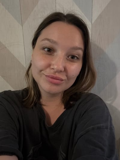 A medium shot shows a woman with brown hair and brown eyes smiling at the camera. She is wearing a dark gray t-shirt. The background is a wall with a diagonal pattern of light and dark gray. There is no information about the location being in the city of Белоус, Russia.